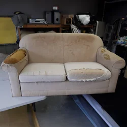 Well-used two-seater sofa in Bexley workshop, fabric worn and cushions damaged, photographed before restoration work begins.