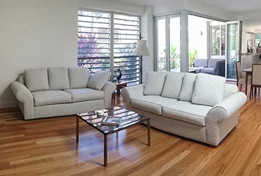 A pair of light-patterned sofas reupholstered in soft neutral fabric, styled in a modern living room with polished timber floors and glass doors opening to a leafy garden.